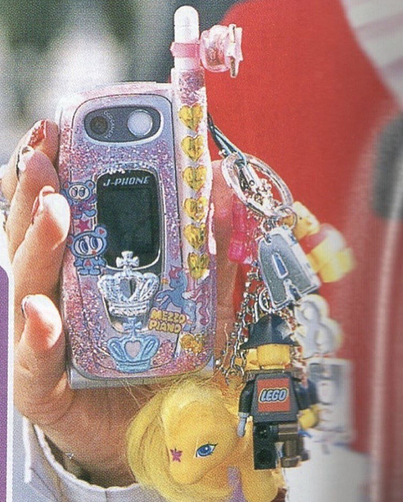 A pink flip phone branded 'J-PHONE', full of charms and stickers, like we used to do in the 2000s.